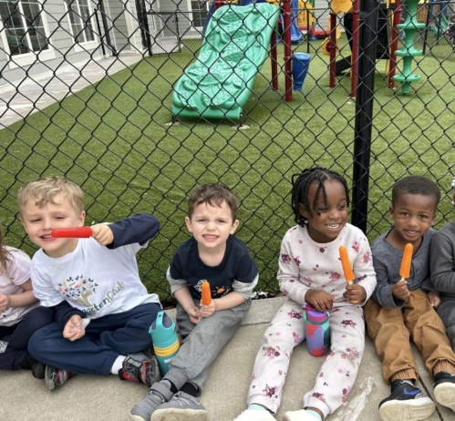 Children sitting outside by playground against a fence eating popsicles and smiling at Cadence academy preschool and daycare 505 murfreesboro
