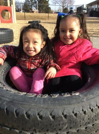 preschool_girls_sitting_in_tire_on_playground_at_cadence_academy_preschool_columbine_littleton_co-333x450