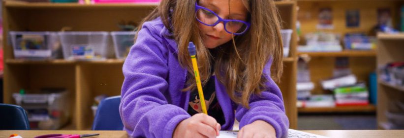 kindergarten to first grade girl in purple jacket and glasses completing an assignment at a table in a classroom