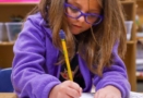 kindergarten to first grade girl in purple jacket and glasses completing an assignment at a table in a classroom