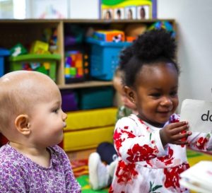 two happy infant girls in photo in infant care classroom with one looking at the other who is reading flashcards with a teacher off camera