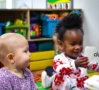 two happy infant girls in photo in infant care classroom with one looking at the other who is reading flashcards with a teacher off camera