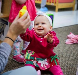 happy infant sitting on a rug playing with a teacher holding a toy while another infant stands in the background