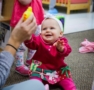 happy infant sitting on a rug playing with a teacher holding a toy while another infant stands in the background