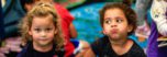 two children in the summer camp program sitting on the carpet, one looking at the camera and one not. the one not is making a silly face both children are having fun
