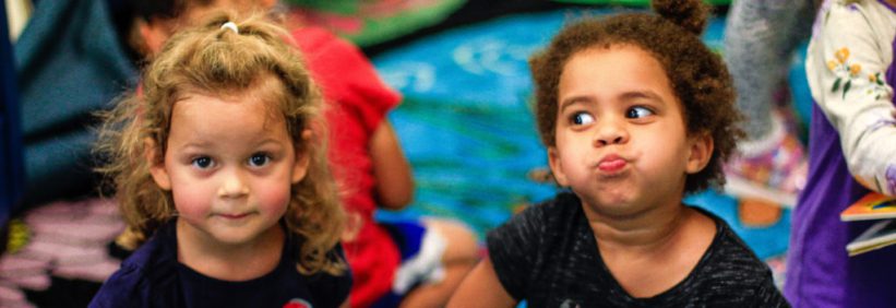 two children in the summer camp program sitting on the carpet, one looking at the camera and one not. the one not is making a silly face both children are having fun