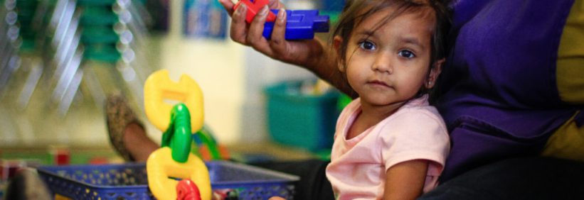 small girl sitting in lap of teacher looking at camera in her childcare program while the teacher shows her different toys