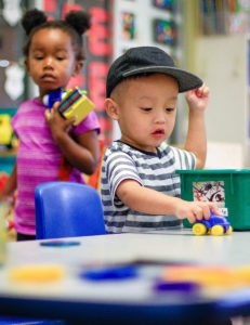 small boy toddler playing with a truck on table with a girl of similar age in daycare carrying toys in background