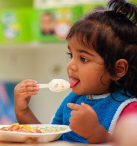Toddler aged child eating lunch provided with tuition by Milestones Academy wearing a small blue apron and sitting at a table